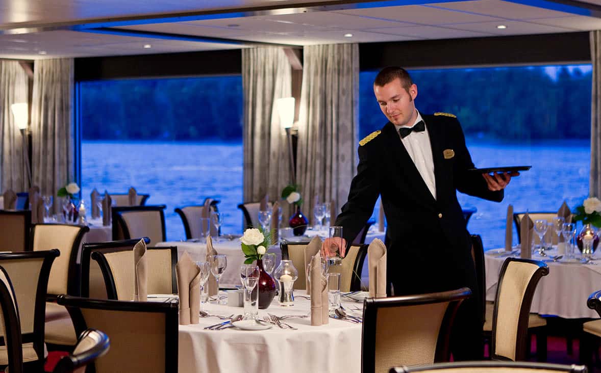 Un serveur en uniforme prépare une salle à manger élégante avec vue sur l’eau, offrant une expérience de voyage de luxe avec TMR Voyages.