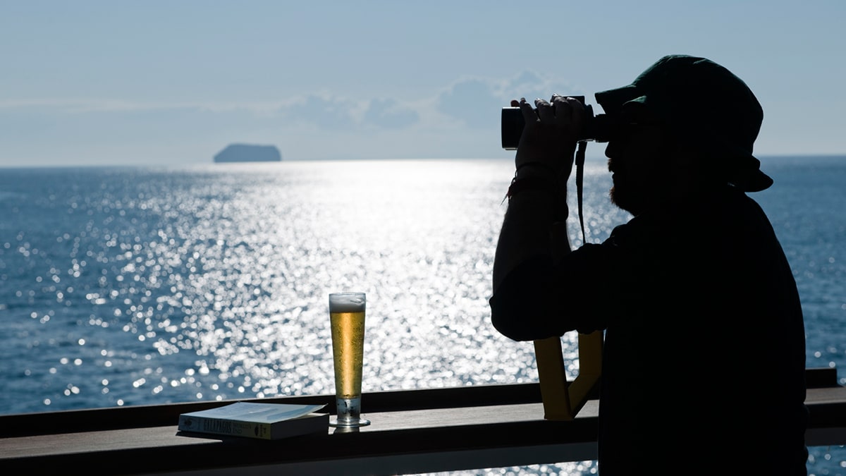 Un voyage d’exception avec TMR Voyages, un homme observant l'horizon depuis un balcon ensoleillé, bière à la main.