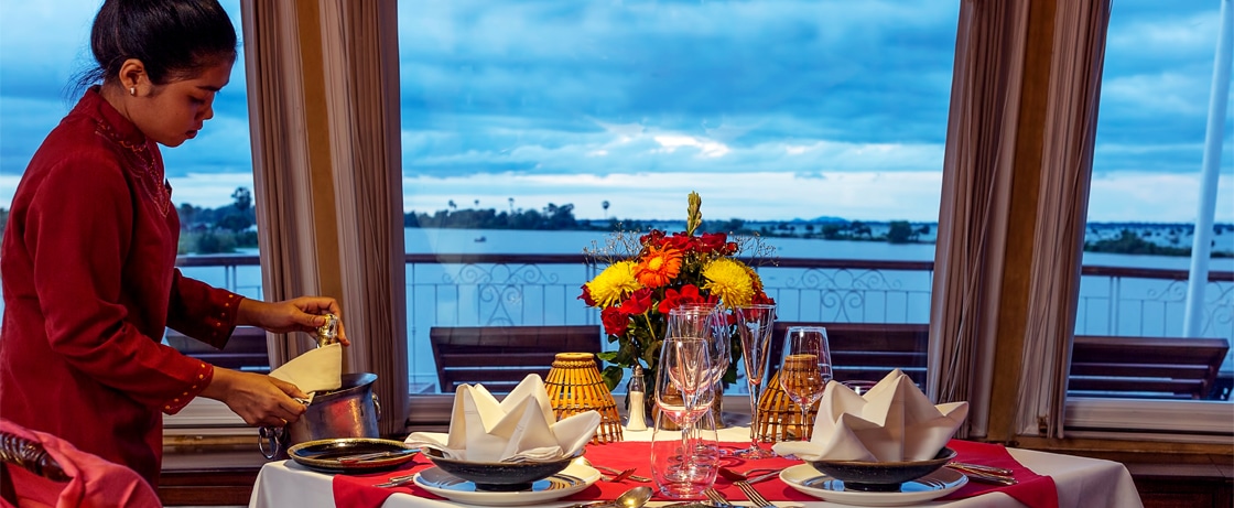Une serveuse prépare une table élégante avec vue sur l'eau, illustrant un voyage de luxe organisé par TMR Voyages.